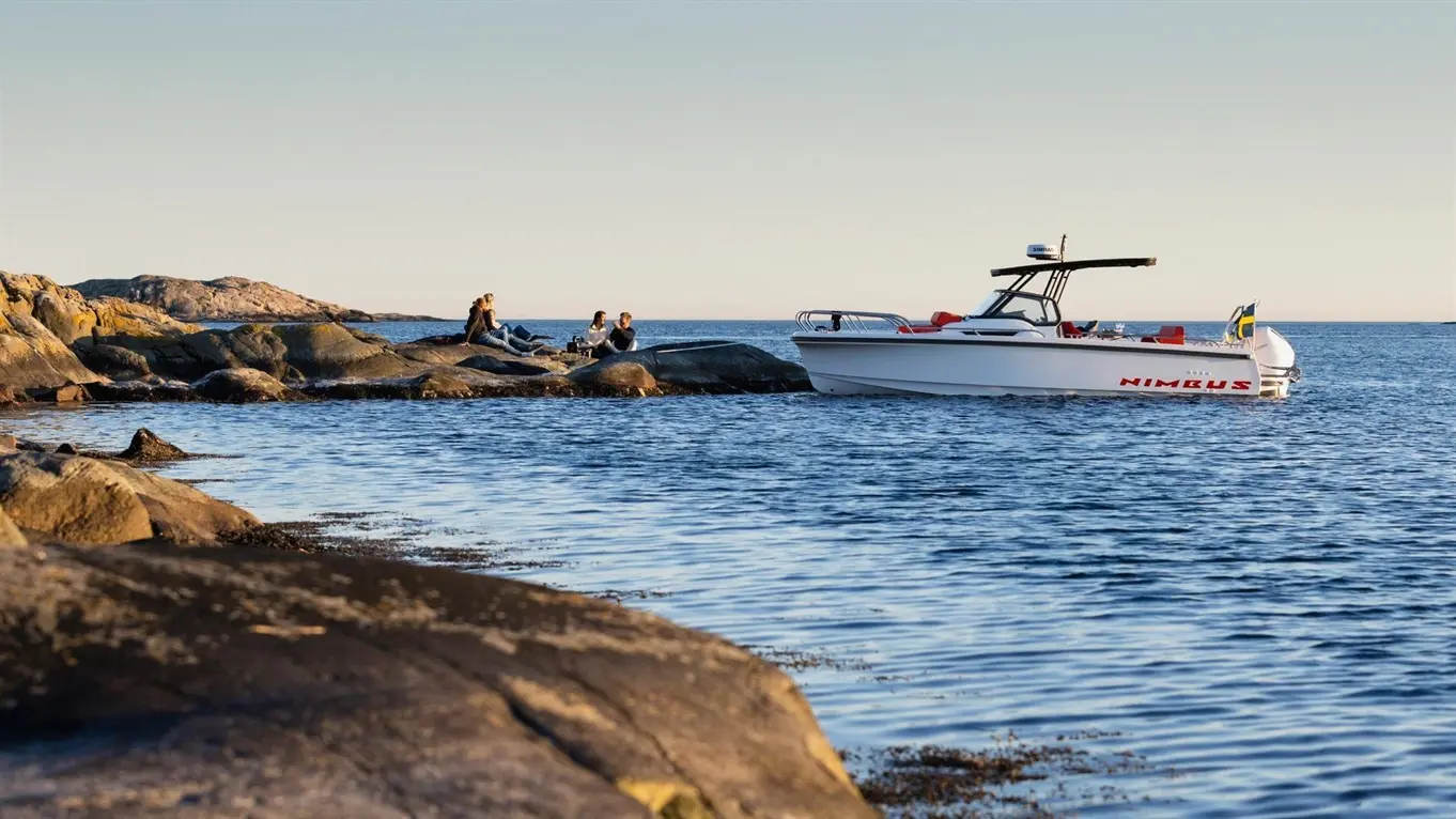 A Nimbus boat anchored near rocky shore where four people relax, enjoying the scenic seaside view at sunset.